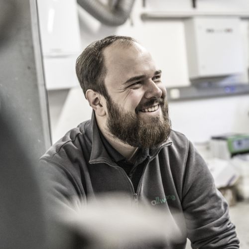 Ein lächelnder Haustechnik-Experte sitzt in einer Werkstatt, umgeben von Heiztechnik-Ausrüstung.