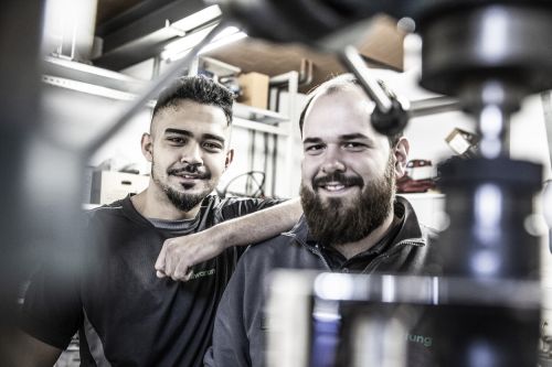 Zwei Männer in Arbeitskleidung lächeln in einer Werkstatt für Heiztechnik und sanitäre Installationen.