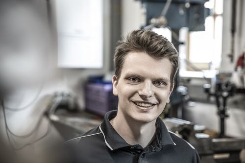 Ein junger Mann lächelt in einer Werkstatt, umgeben von moderner Heiztechnik und Klimatechnik.