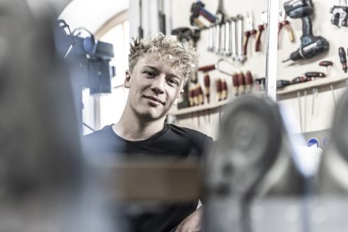 Ein junger Mann lächelt in einer Werkstatt, umgeben von Werkzeugen für Heiztechnik und Klimatechnik.