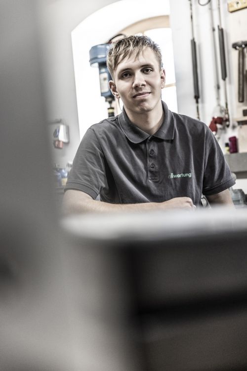 Ein junger Mann in einem grauen Poloshirt sitzt lächelnd in einer Werkstatt für Klimatechnik.