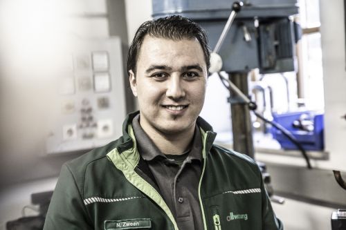 Ein Monteur in Arbeitskleidung lächelt in einer Werkstatt, spezialisiert auf Heiztechnik und Allwartung.