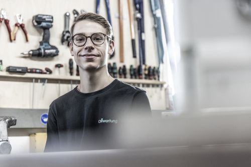 Ein junger Mann lächelt in einer Werkstatt, umgeben von Werkzeugen für Klimatechnik an einer Wand.