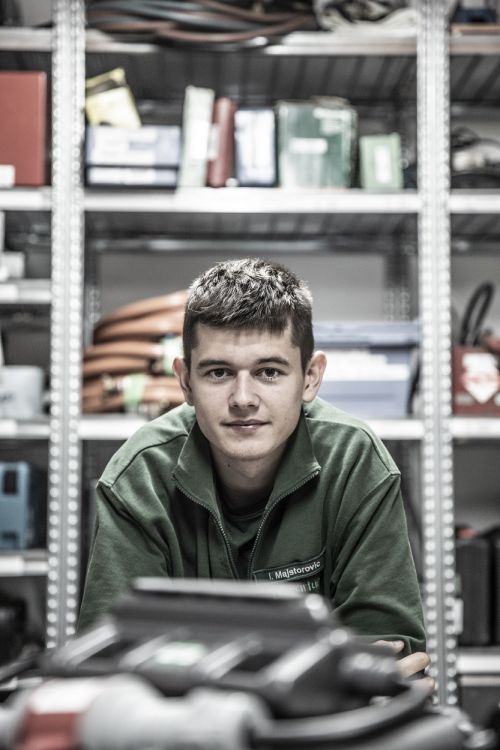 Ein junger Mann in Arbeitskleidung lehnt sich in einer Werkstatt auf ein Regal, umgeben von Werkzeugen für Klimatechnik.