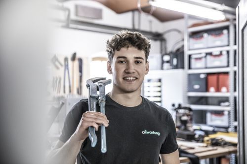 Ein junger Mann steht lächelnd in einer Werkstatt und hält Werkzeuge, bereit für Arbeiten in der Klimatechnik.