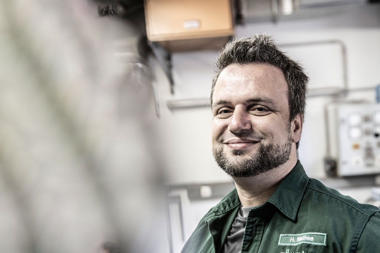 Ein lächelnder Mann in grüner Arbeitskleidung steht in einem Raum voller Heiztechnik und lüftungstechnischer Anlagen.