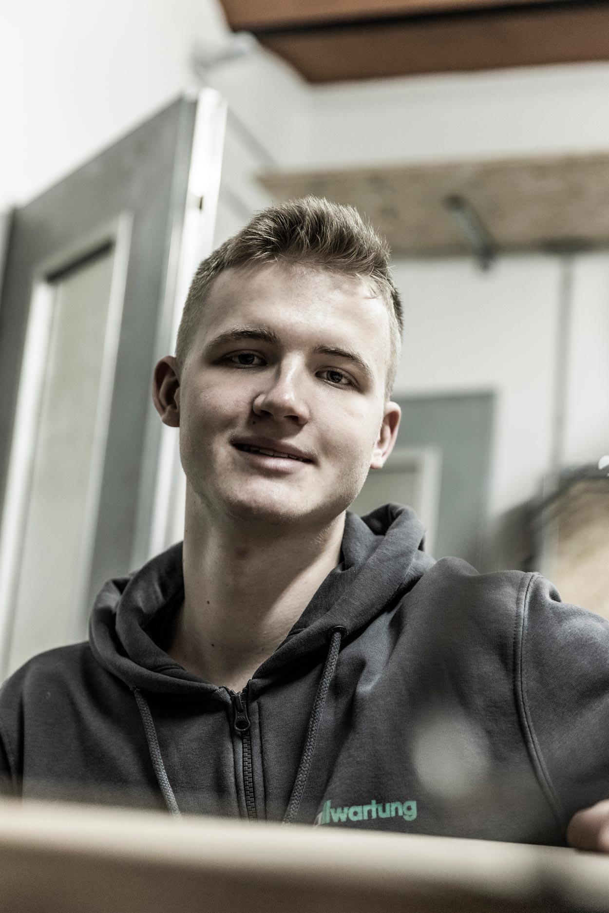 Ein junger Mann in Arbeitskleidung lächelt in einer Werkstatt, die auf Klimatechnik spezialisiert ist.